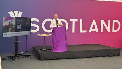 Jim Fairlie speaks on a stage with a Scottish Government background.